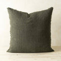The Indaba Trading 24x24 Lina Linen Pillow Avocado, a square dark gray-green cover with filler and frayed edge, sits on a light wooden surface against an off-white wall, crafted from sustainable materials for timeless style.