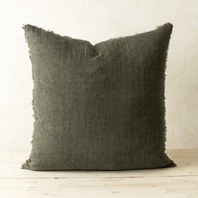 The Indaba Trading 24x24 Lina Linen Pillow Avocado, a square dark gray-green cover with filler and frayed edge, sits on a light wooden surface against an off-white wall, crafted from sustainable materials for timeless style.