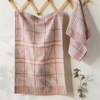 Two Bisque Finley Check Dishtowels Set of 2 by Danica Heirloom hang on wooden hooks against a white wall—one large and draped, the other small and folded. A eucalyptus sprig accents the corner.