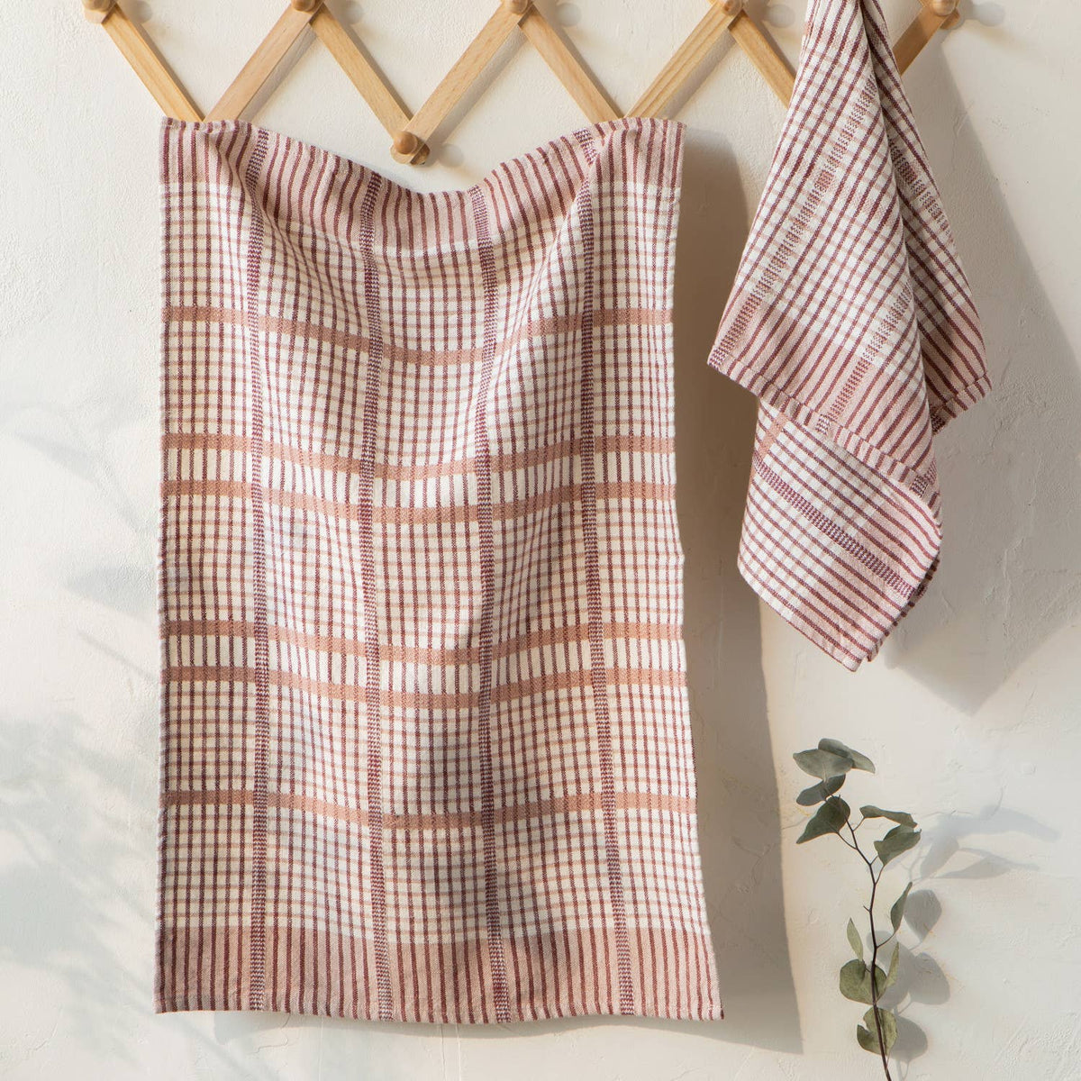 Two Bisque Finley Check Dishtowels Set of 2 by Danica Heirloom hang on wooden hooks against a white wall—one large and draped, the other small and folded. A eucalyptus sprig accents the corner.