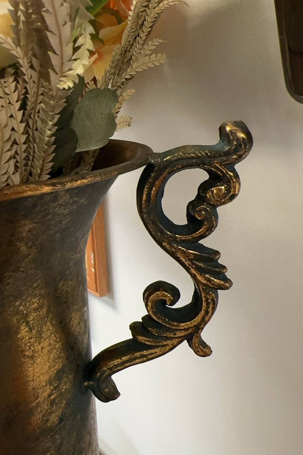 A close-up of the Vagabond Vintage Gold Trophy Vase with Handles showcases its ornate, antique gold handle with intricate scrollwork, attached to the vase and filled with dried foliage.