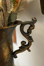 A close-up of the Vagabond Vintage Gold Trophy Vase with Handles showcases its ornate, antique gold handle with intricate scrollwork, attached to the vase and filled with dried foliage.