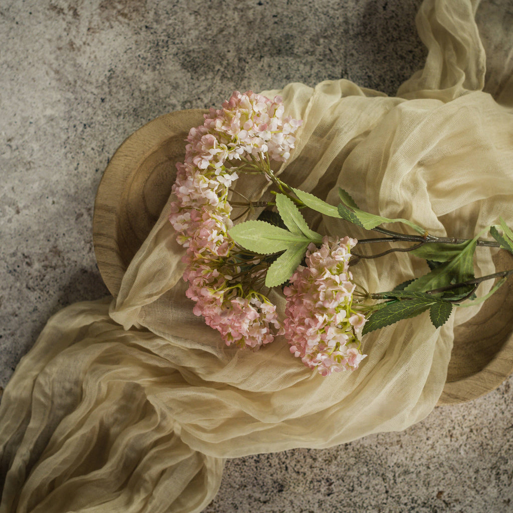 The 25" PINK FAUX HYDRANGEA SPRAY by Ragon House, with green leaves, rests on a round wooden tray draped in sheer beige fabric atop a textured stone surface.