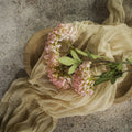 The 25" PINK FAUX HYDRANGEA SPRAY by Ragon House, with green leaves, rests on a round wooden tray draped in sheer beige fabric atop a textured stone surface.