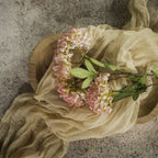The 25" PINK FAUX HYDRANGEA SPRAY by Ragon House, with green leaves, rests on a round wooden tray draped in sheer beige fabric atop a textured stone surface.