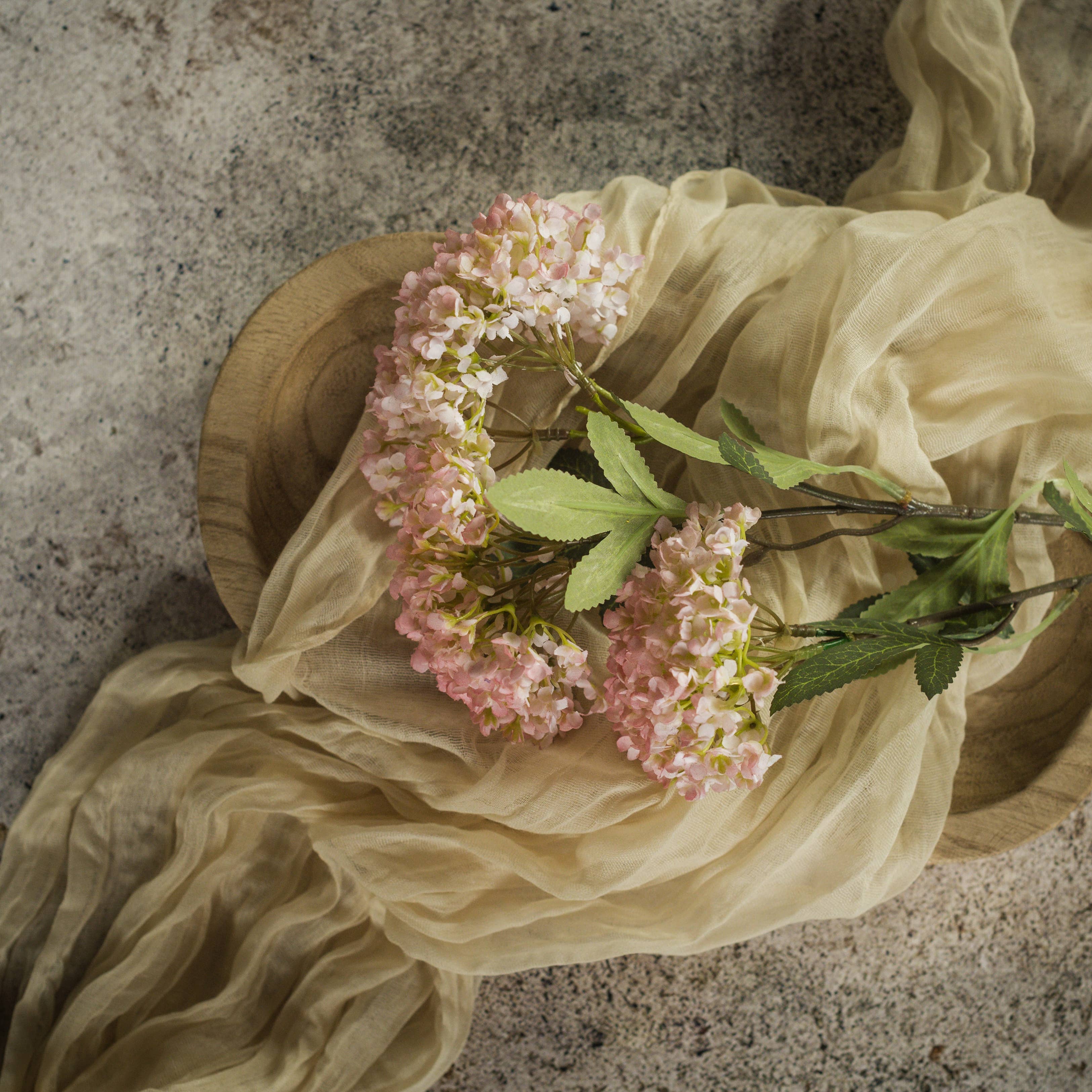 The 25" PINK FAUX HYDRANGEA SPRAY by Ragon House, with green leaves, rests on a round wooden tray draped in sheer beige fabric atop a textured stone surface.