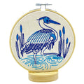 The Blue Heron Complete Embroidery Kit by Hook, Line & Tinker features a blue heron stitched in blue thread on cream fabric with water and reeds, displayed in a wooden hoop with stand.