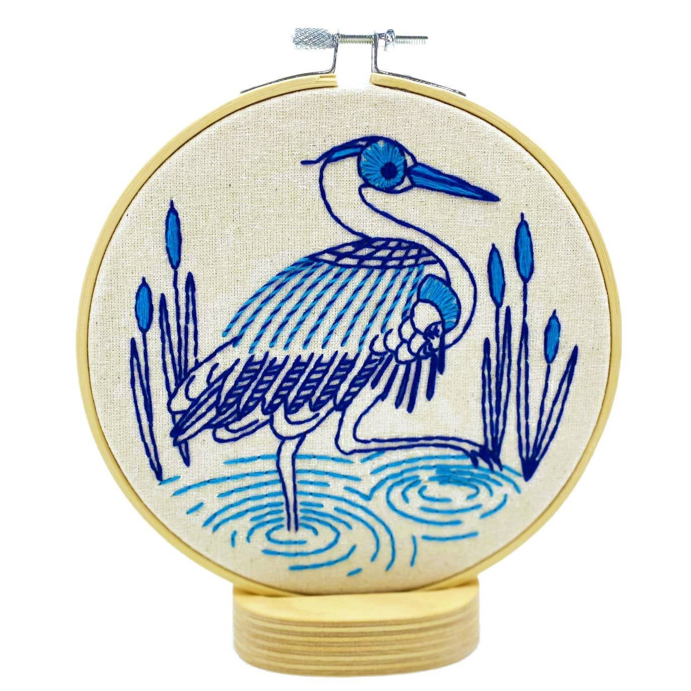 The Blue Heron Complete Embroidery Kit by Hook, Line & Tinker features a blue heron stitched in blue thread on cream fabric with water and reeds, displayed in a wooden hoop with stand.