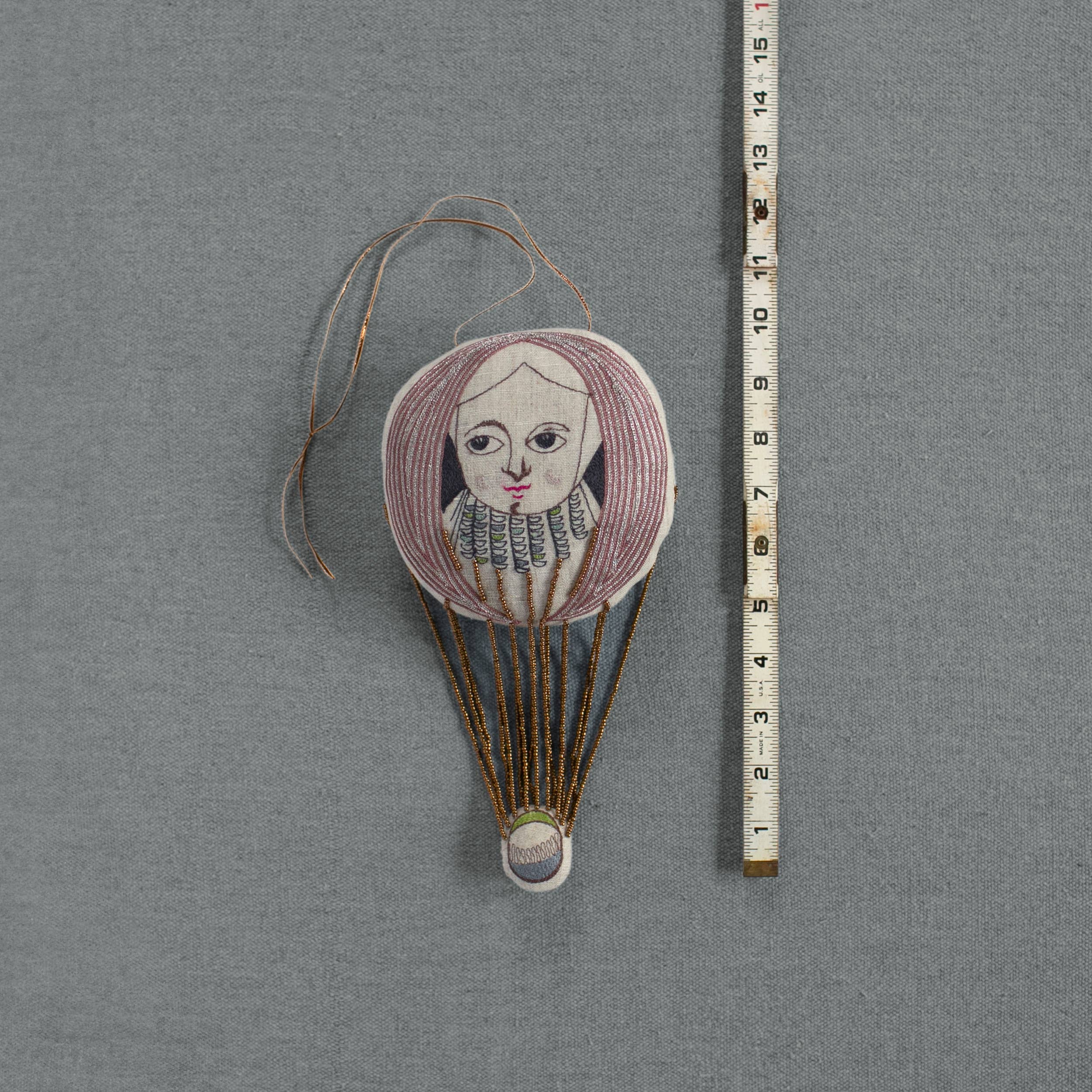 The Wonderful Wizard / Balloon by Skippy Cotton is an oval embroidered ornament featuring a hot air balloon with a woman's pink-haired face, measuring 13 cm—a unique Oz-inspired textile wallhanging on gray fabric.