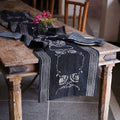 A rustic wooden table features the Ichcha Deepest Navy & White Table Linens Cotton Table Runner - Danya, folded napkins, and a small glass vase with pink flowers at the center, surrounded by wooden chairs.