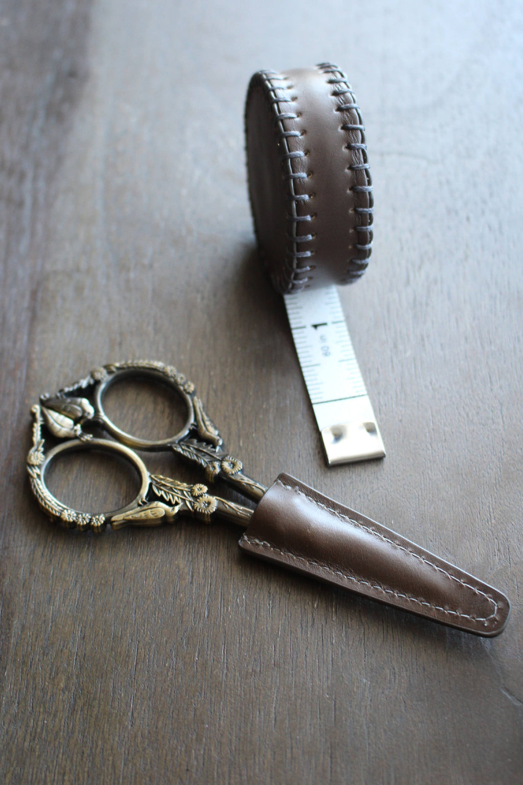 NNK Press Leather Scissor Sheath, shown with ornate embroidery scissors and a round brown leather tape measure, is displayed on a wooden surface. Perfect as a scissors protector.