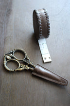 NNK Press Leather Scissor Sheath, shown with ornate embroidery scissors and a round brown leather tape measure, is displayed on a wooden surface. Perfect as a scissors protector.