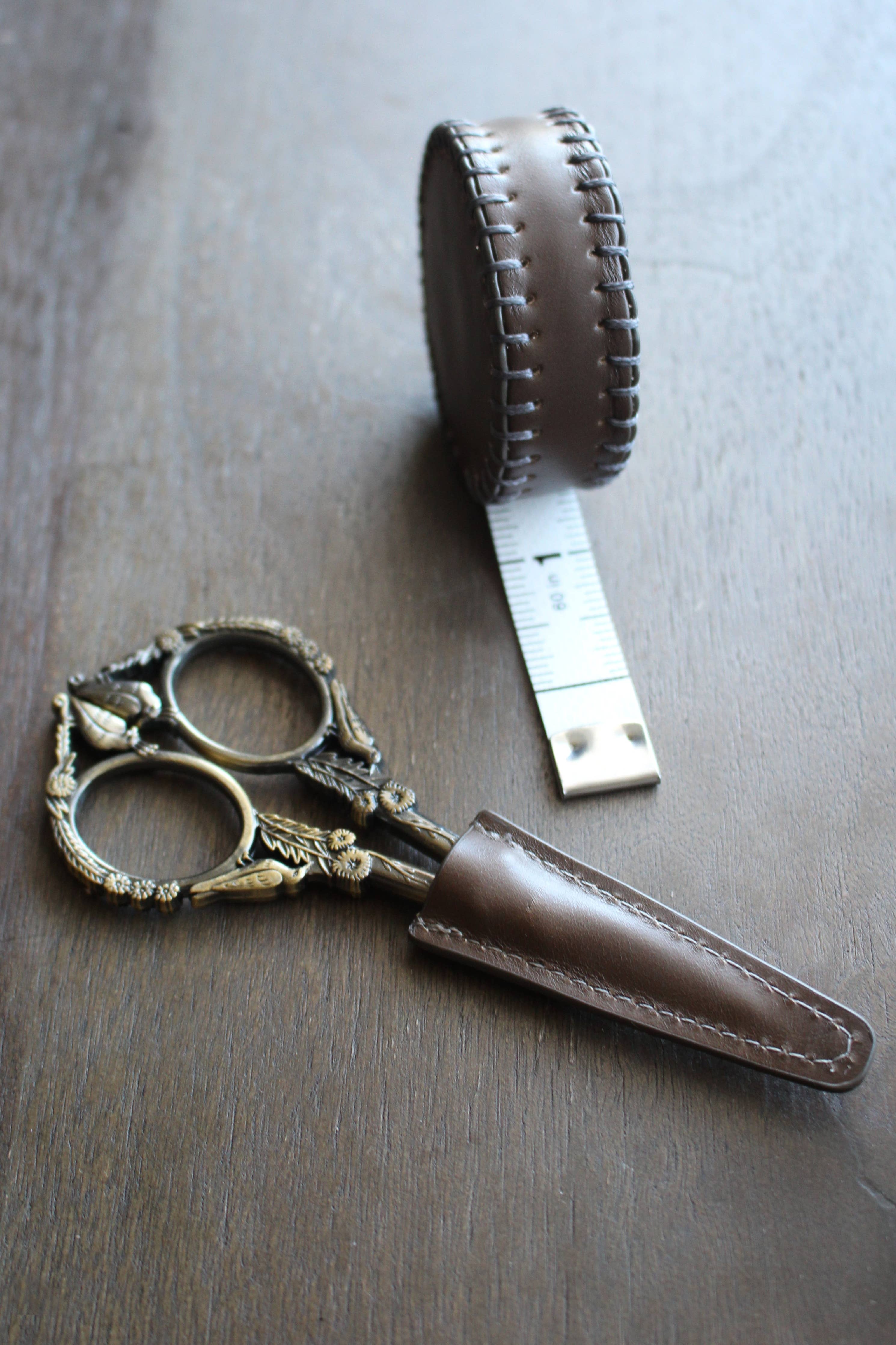 NNK Press Leather Scissor Sheath, shown with ornate embroidery scissors and a round brown leather tape measure, is displayed on a wooden surface. Perfect as a scissors protector.