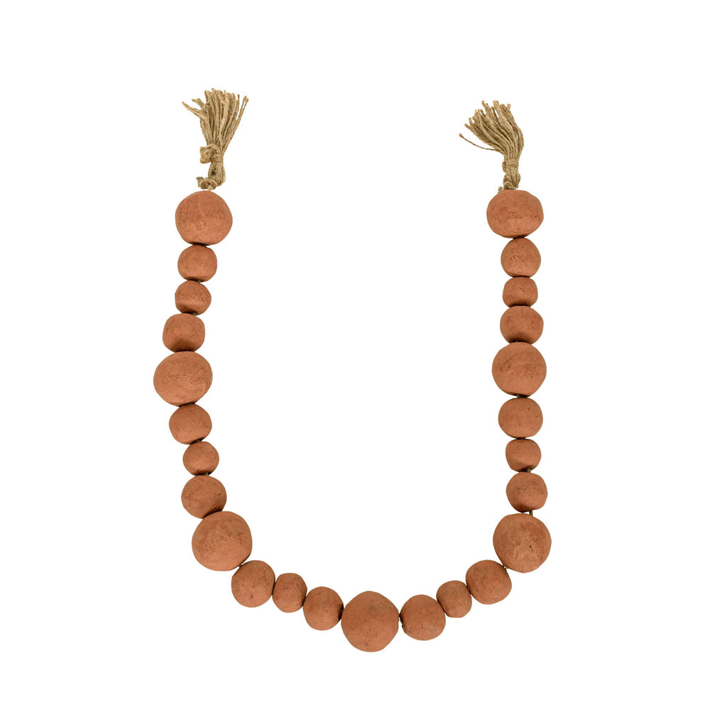 The Foreside Home & Garden Clay Garland Terracotta + Jute features hand-formed round reddish-brown clay beads in varying sizes, arranged in a U-shape with tan jute tassels tied at each end.