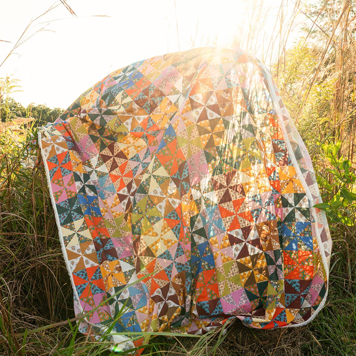 A colorful patchwork quilt with a pinwheel pattern is draped over tall grass, sunlight shining through, with the Handker Bandanas Calico Quilted Tote resting on the golden grass in the background.