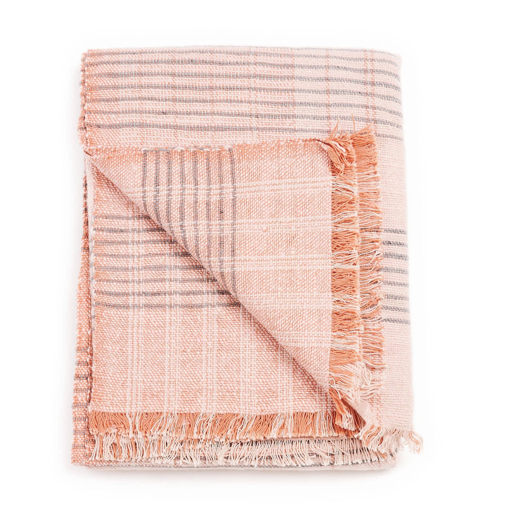A folded Silica Rose Throw by Variously, crafted in merino wool in shades of salmon with a plaid pattern of thin gray stripes and finished with fringed edges.