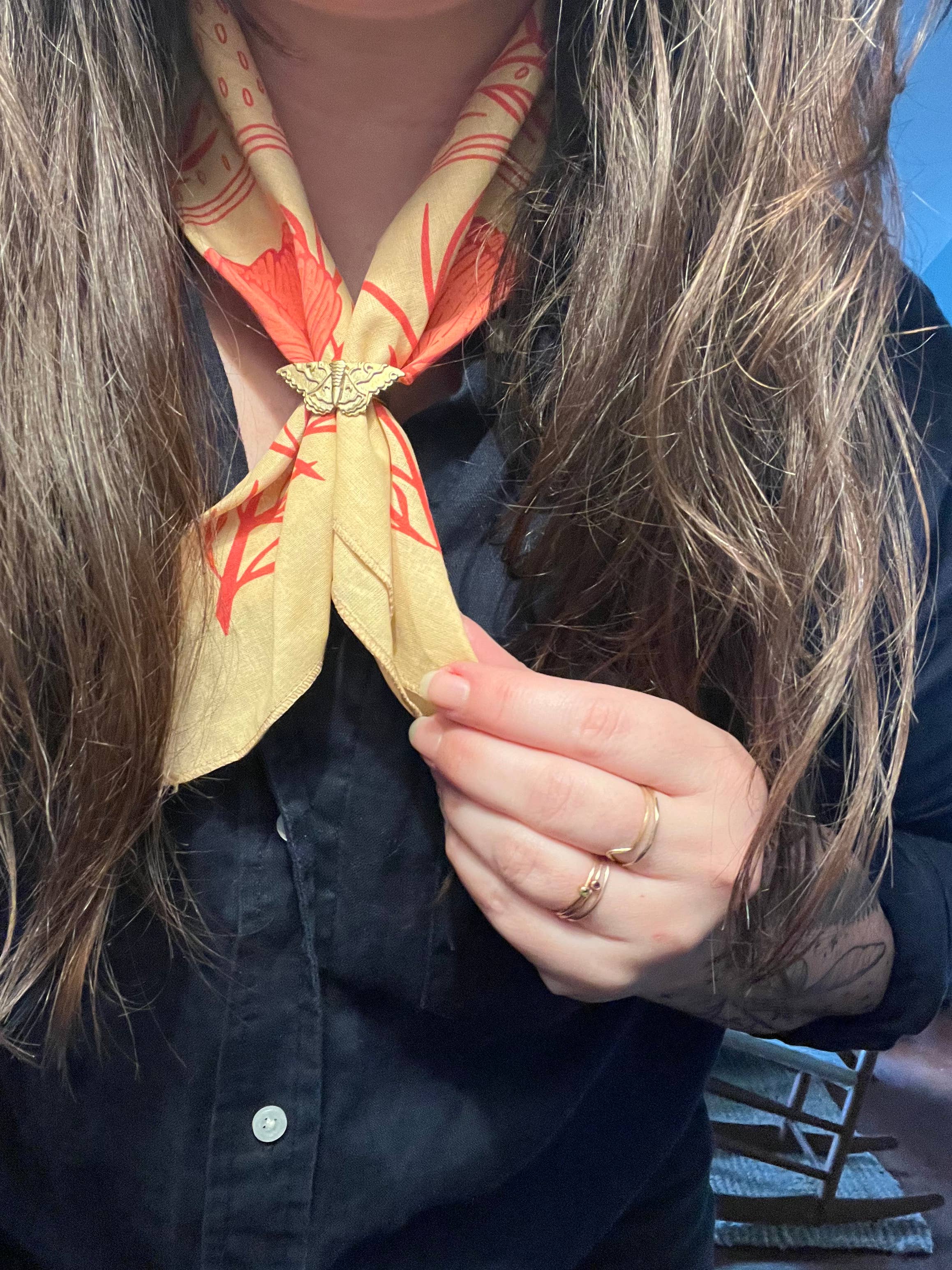A person with long brown hair and gold rings fastens the Pen+Pine Pink Cosmos Bandana, an eco-friendly, silk screened cotton accessory, around their neck. They wear a dark button-up shirt and use a butterfly-shaped pin.
