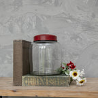 The 8" CLEAR GLASS JAR W/ RED LID by Ragon House sits between two vintage books, with artificial red and white flowers nearby on a wooden surface against a textured gray wall.