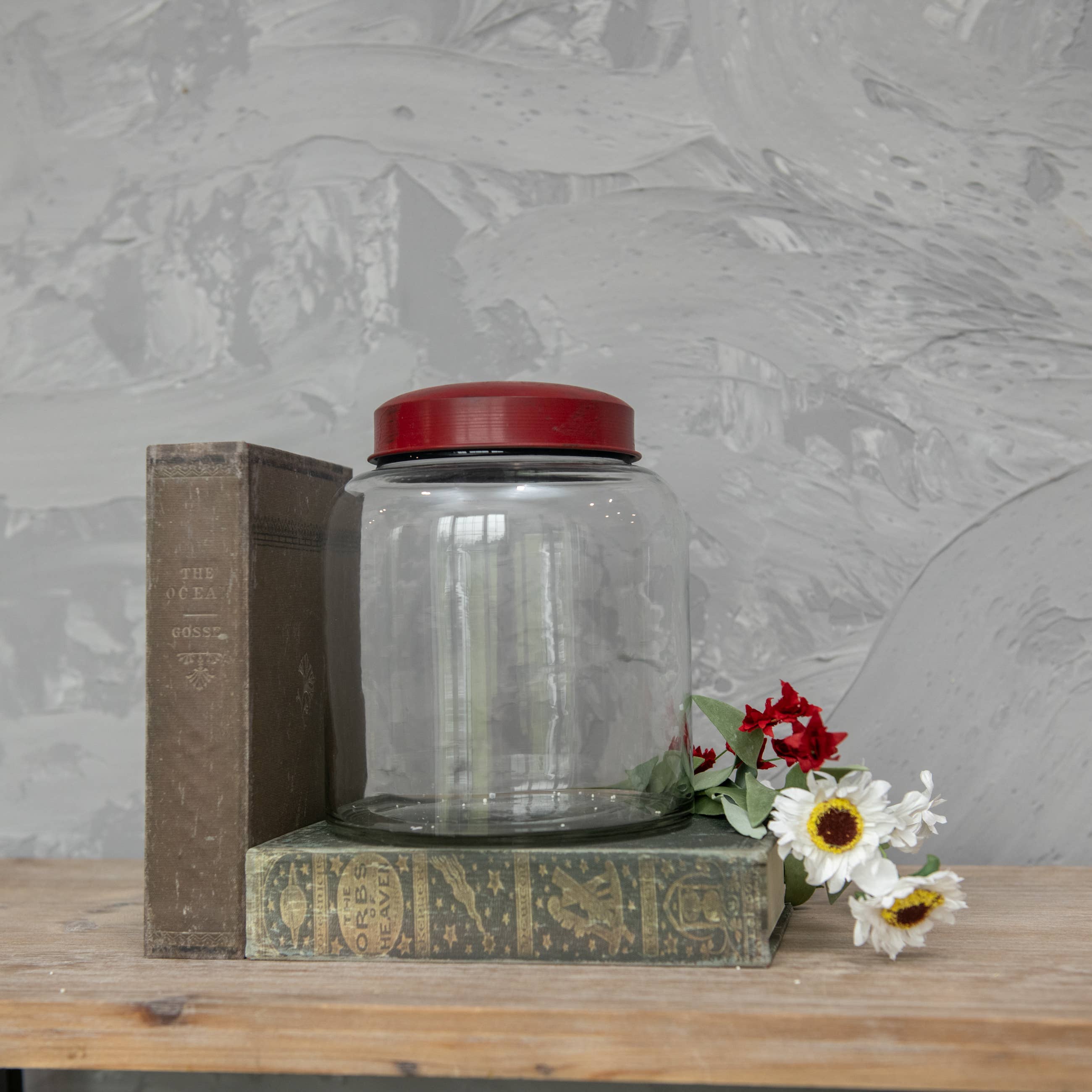 The 8" CLEAR GLASS JAR W/ RED LID by Ragon House sits between two vintage books, with artificial red and white flowers nearby on a wooden surface against a textured gray wall.