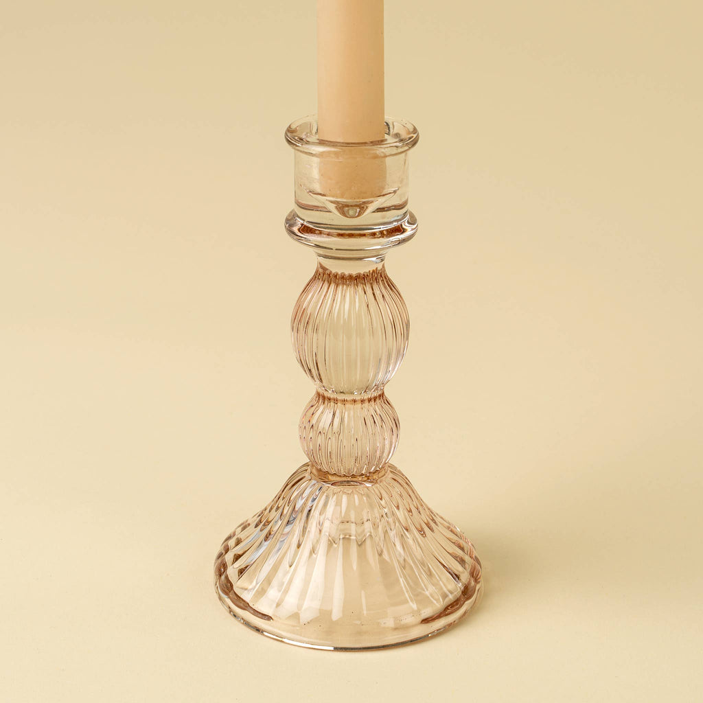 The Indaba Trading Callista Glass Candleholder Large in blush, paired with a beige taper candle, is perfect for vintage-inspired decor and displayed against a light beige background.