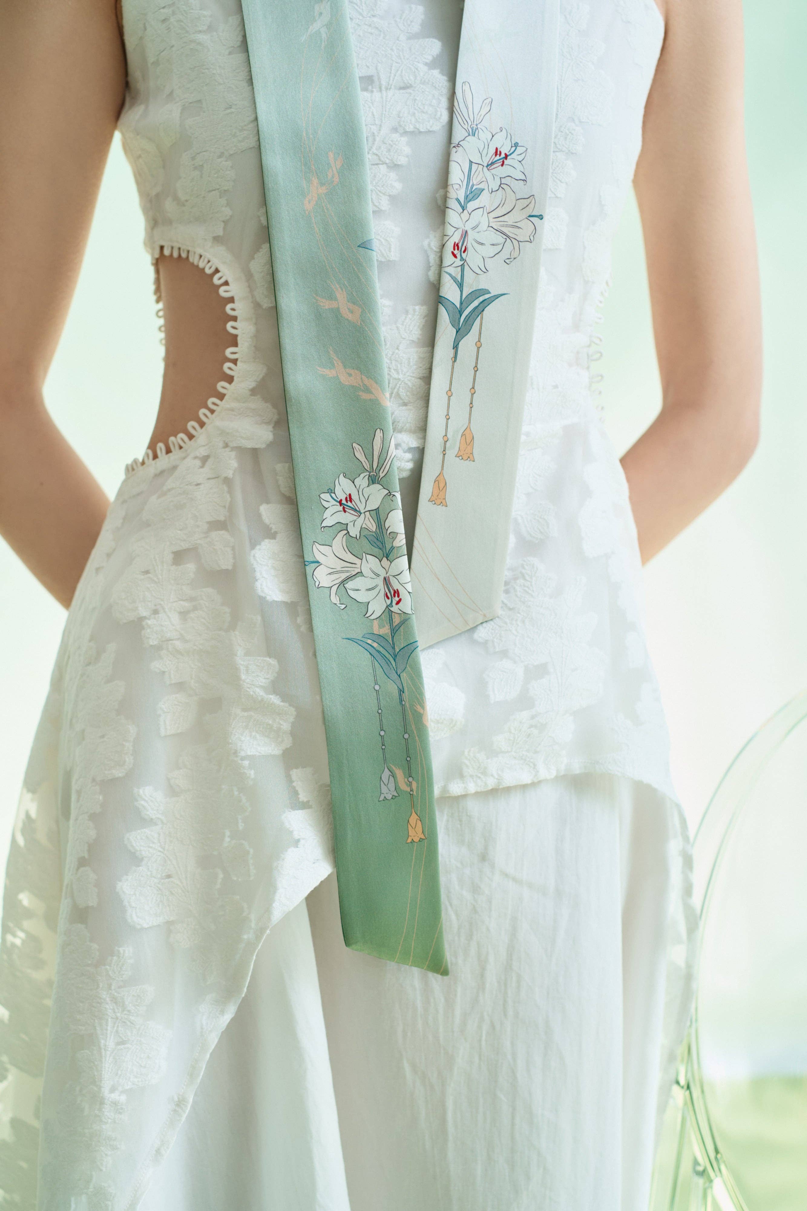 A person wears a sleeveless white dress with floral cut-outs, paired with the Lost Pattern NYC Lily Silk Twilly Scarf in green, featuring delicate florals. A softly lit, pastel background adds grace to the scene.