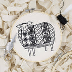 The Sweater Weather Sheep Complete Embroidery Kit by Hook, Line & Tinker features a hoop holding fabric with a black-stitched sheep in a patterned sweater, plus scissors and thread on cream-colored strips.