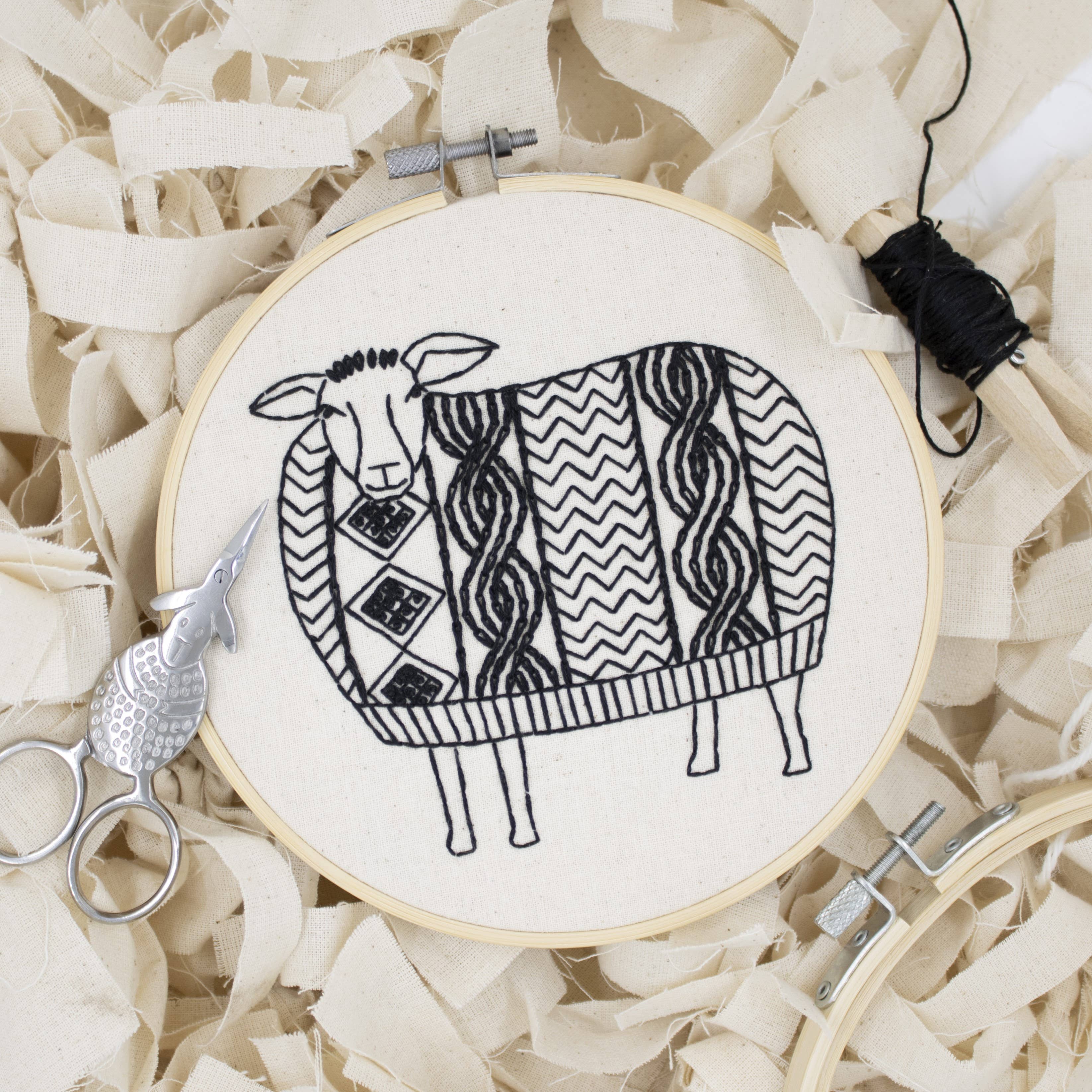 The Sweater Weather Sheep Complete Embroidery Kit by Hook, Line & Tinker features a hoop holding fabric with a black-stitched sheep in a patterned sweater, plus scissors and thread on cream-colored strips.