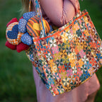 Dressed in pink, a person carries the Calico Quilted Tote by Handker Bandanas—a colorful bandana cotton bag with a patchwork triangle design, holding two fabric balls against a softly blurred grassy background.