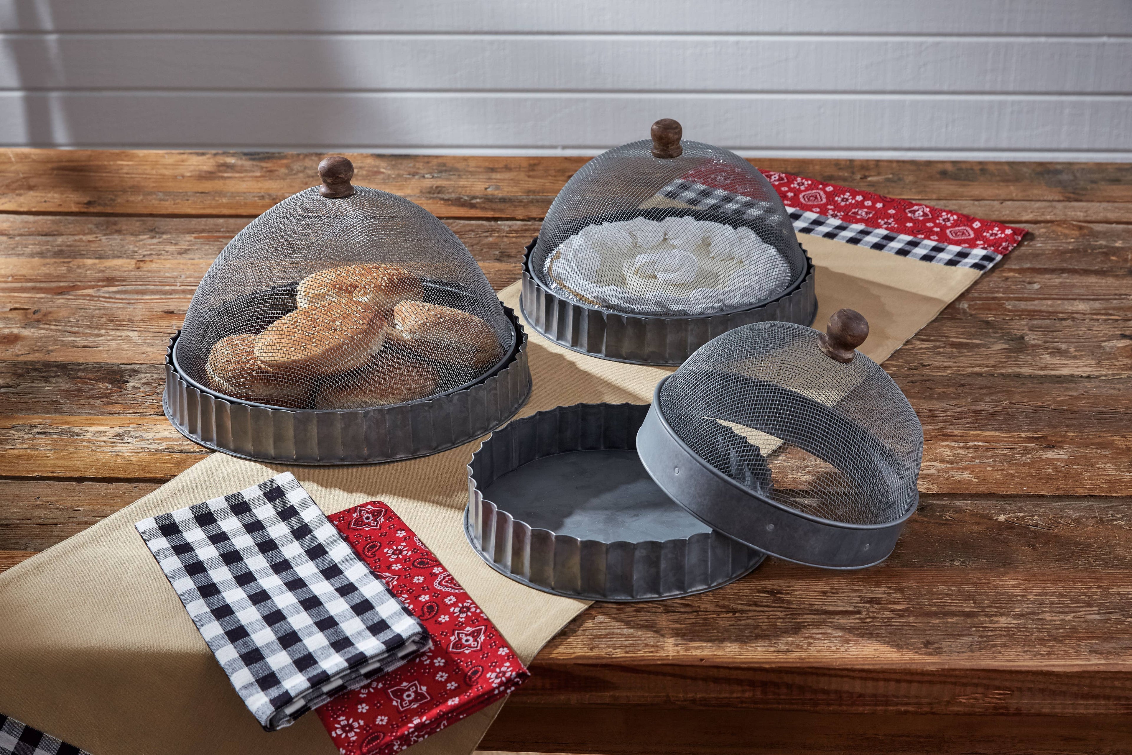 The Park Designs Covington Screen Covers with Bases (Set of 3) display bread, white napkins, and offer an empty tray on a wooden table, each protected by a mesh dome; checkered and red fabric napkins are folded nearby.