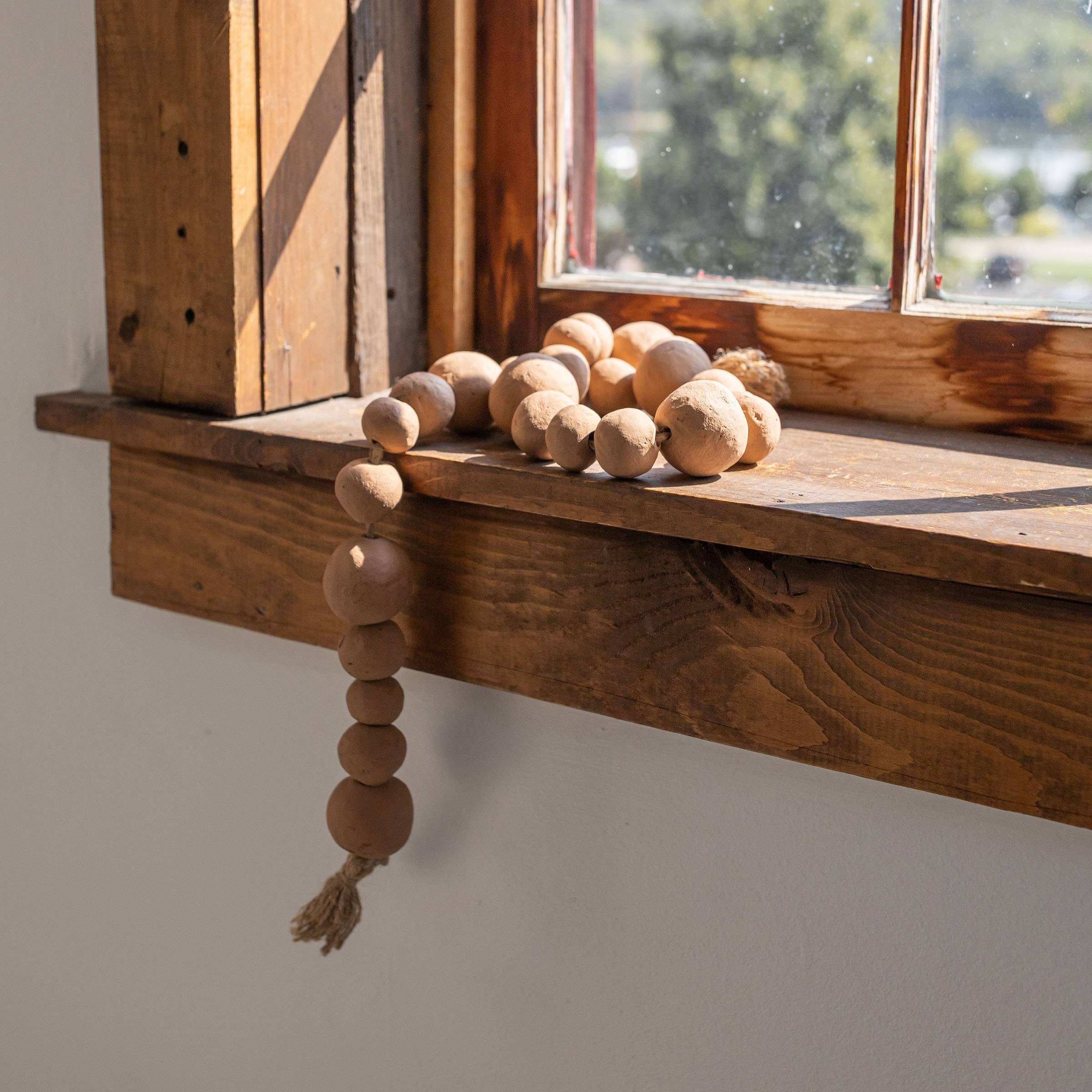 The Clay Garland Terracotta + Jute by Foreside Home & Garden, with hand-formed beads and a tassel, is draped across a rustic wooden windowsill as sunlight streams through, casting elegant shadows on the wood.