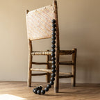 A rustic wooden chair with a woven seat and backrest sits on a wooden floor, draped with Foreside Home & Garden's Black Terracotta Bead + Jute Garland, its large beads cascading nearly to the floor.