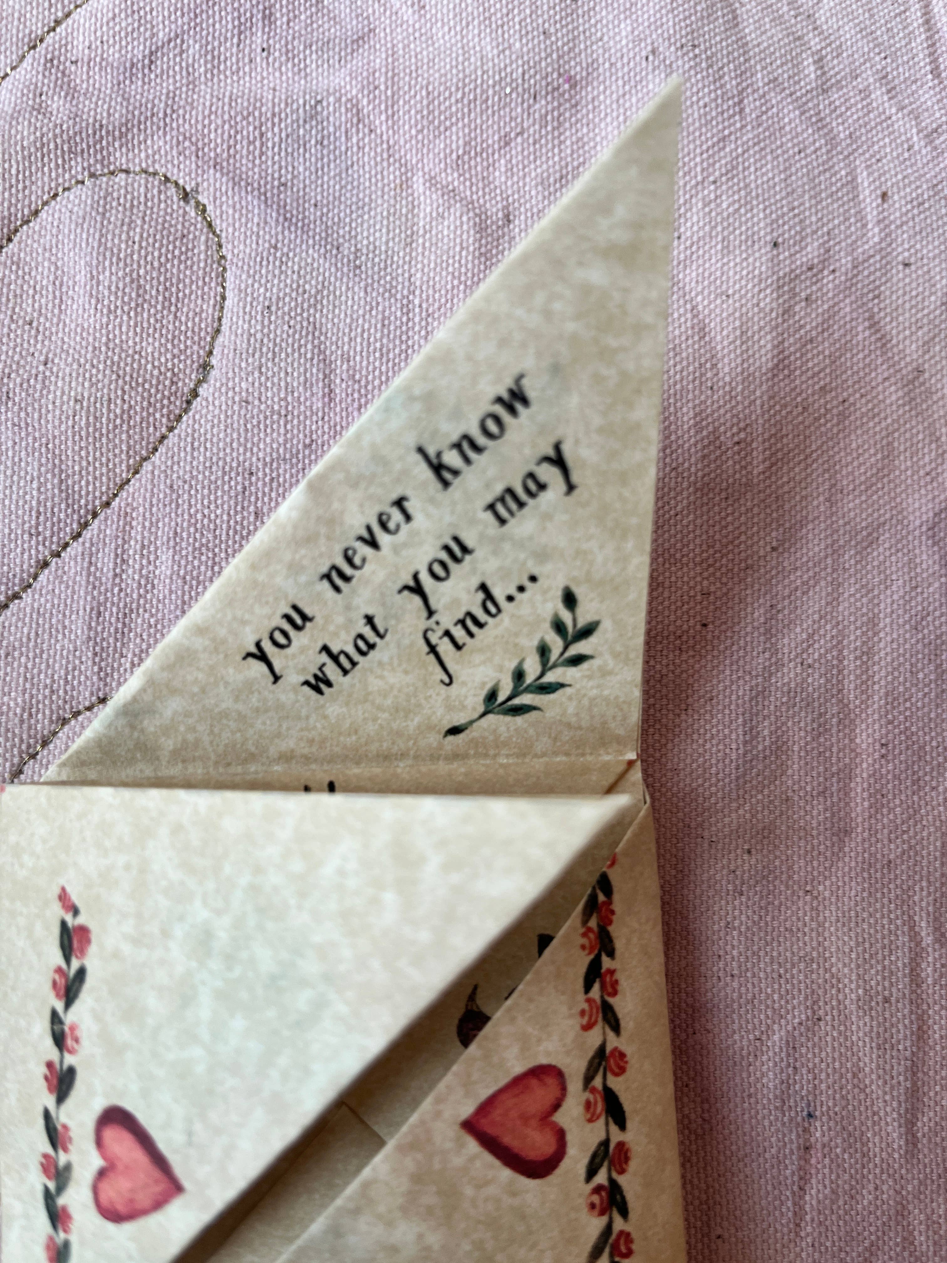 A folded creamy white cardstock envelope from Parcel’s Victorian Valentine Puzzle: Romantic rests on pink fabric with heart and leaf details, featuring the text “you never know what you may find...” on one flap.