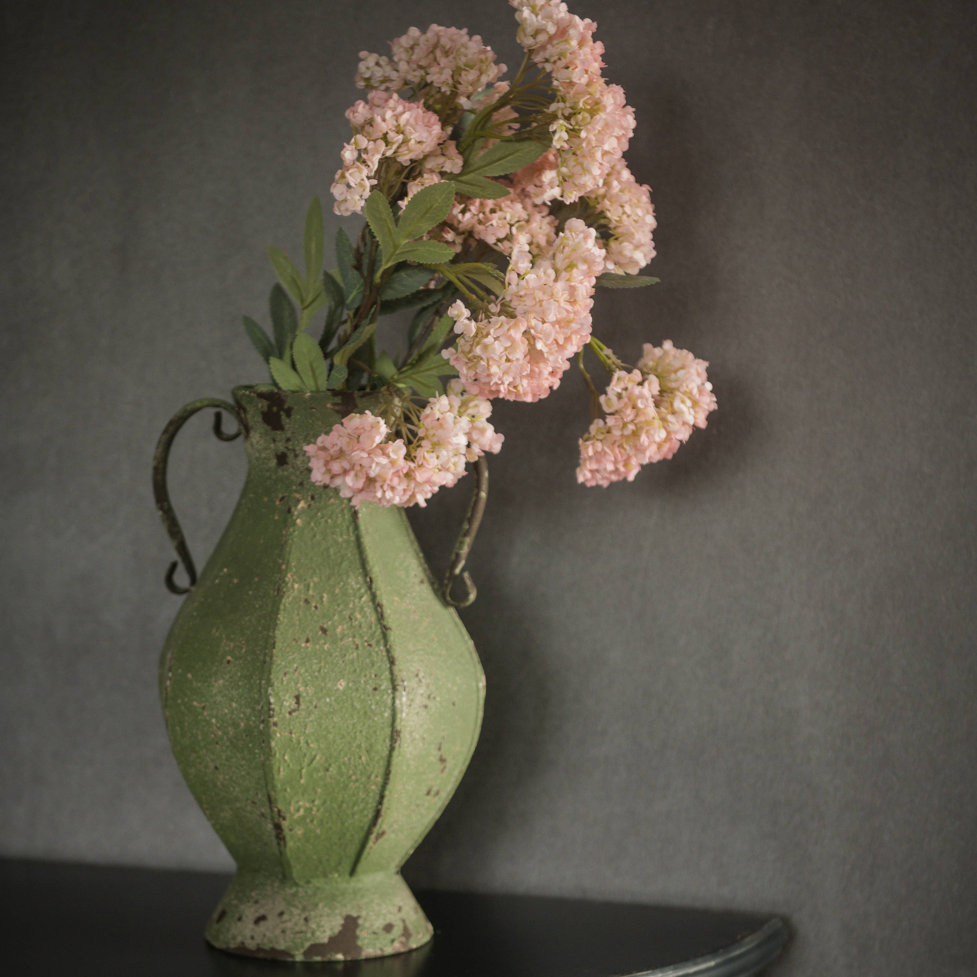 A weathered green metal vase with handles holds a 25" Pink Faux Hydrangea Spray from Ragon House, set against a plain dark gray background on a black surface.
