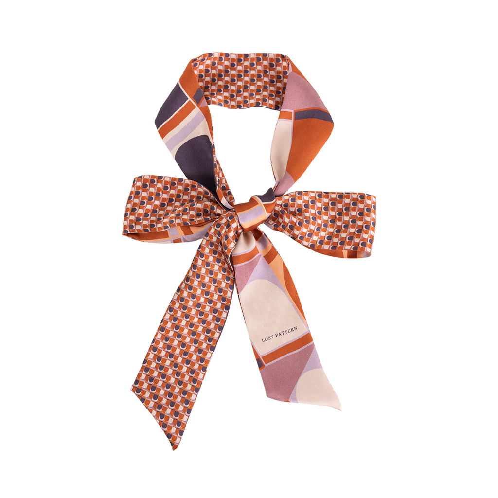 The Silk Skinny Scarf - Clay by Lost Pattern NYC is a double-sided silk satin piece with geometric patterns in orange, purple, and pink—perfect as a chic hair accessory or tied in a bow against a white background.