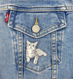 A close-up of a blue denim jacket pocket with a silver button and red Levi's tag, featuring the Wholesale Applique Gray Tabby Cat, Kitten, Pets, Animals Iron on Patch lying down on the pocket.