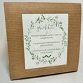 A Grief Kit from Lonely Worm Farm—a brown box with a white “grief kit” label, encircled by a green floral wreath. The label details that it includes directions and ecologically grown herbs for making a grief tincture, offering a meaningful remembrance gift to honor loved ones.