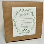 A Grief Kit from Lonely Worm Farm—a brown box with a white “grief kit” label, encircled by a green floral wreath. The label details that it includes directions and ecologically grown herbs for making a grief tincture, offering a meaningful remembrance gift to honor loved ones.