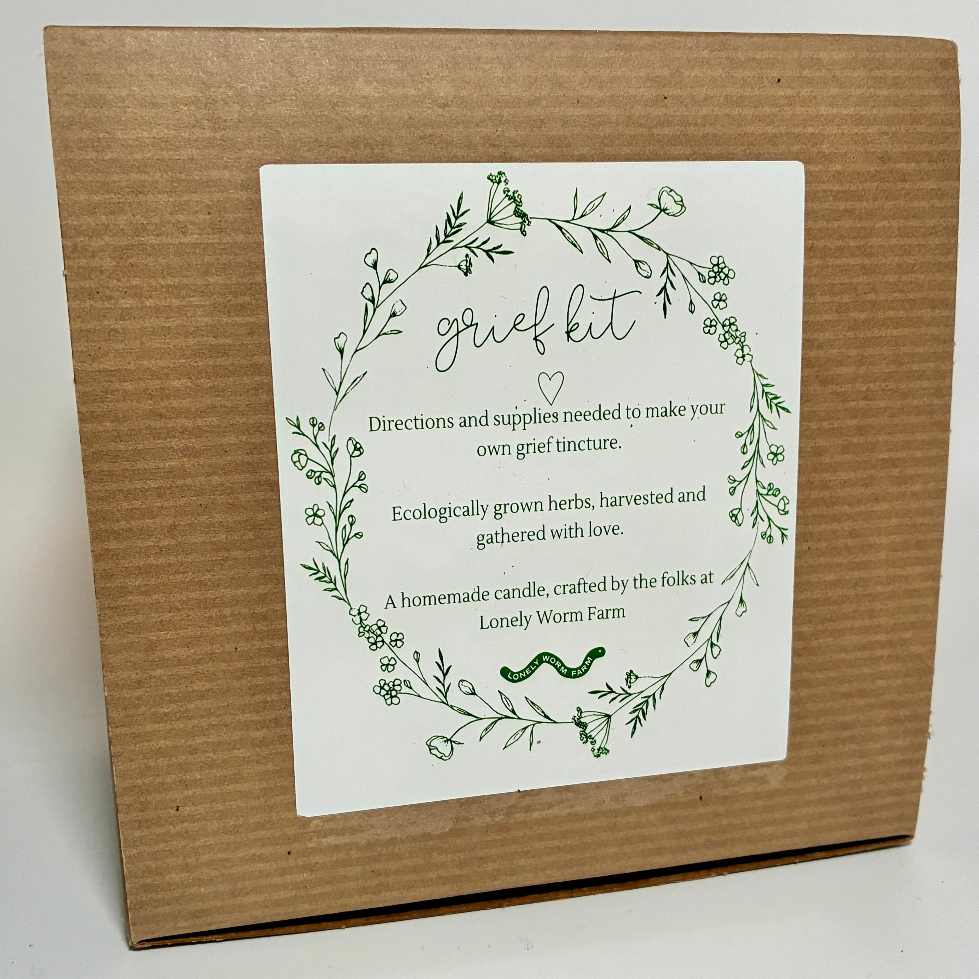 A Grief Kit from Lonely Worm Farm—a brown box with a white “grief kit” label, encircled by a green floral wreath. The label details that it includes directions and ecologically grown herbs for making a grief tincture, offering a meaningful remembrance gift to honor loved ones.