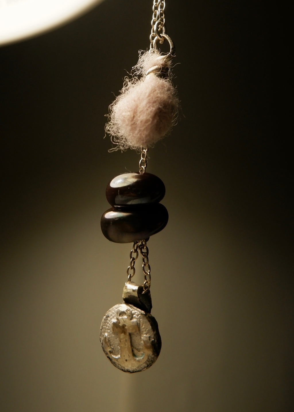 A close-up of the Sunk • Ash Espinoza x Thyne Studio necklace by Thyne Goods, featuring a round textured charm, stacked dark beads, and a small wool ball on a recycled sterling silver chain against a neutral background.