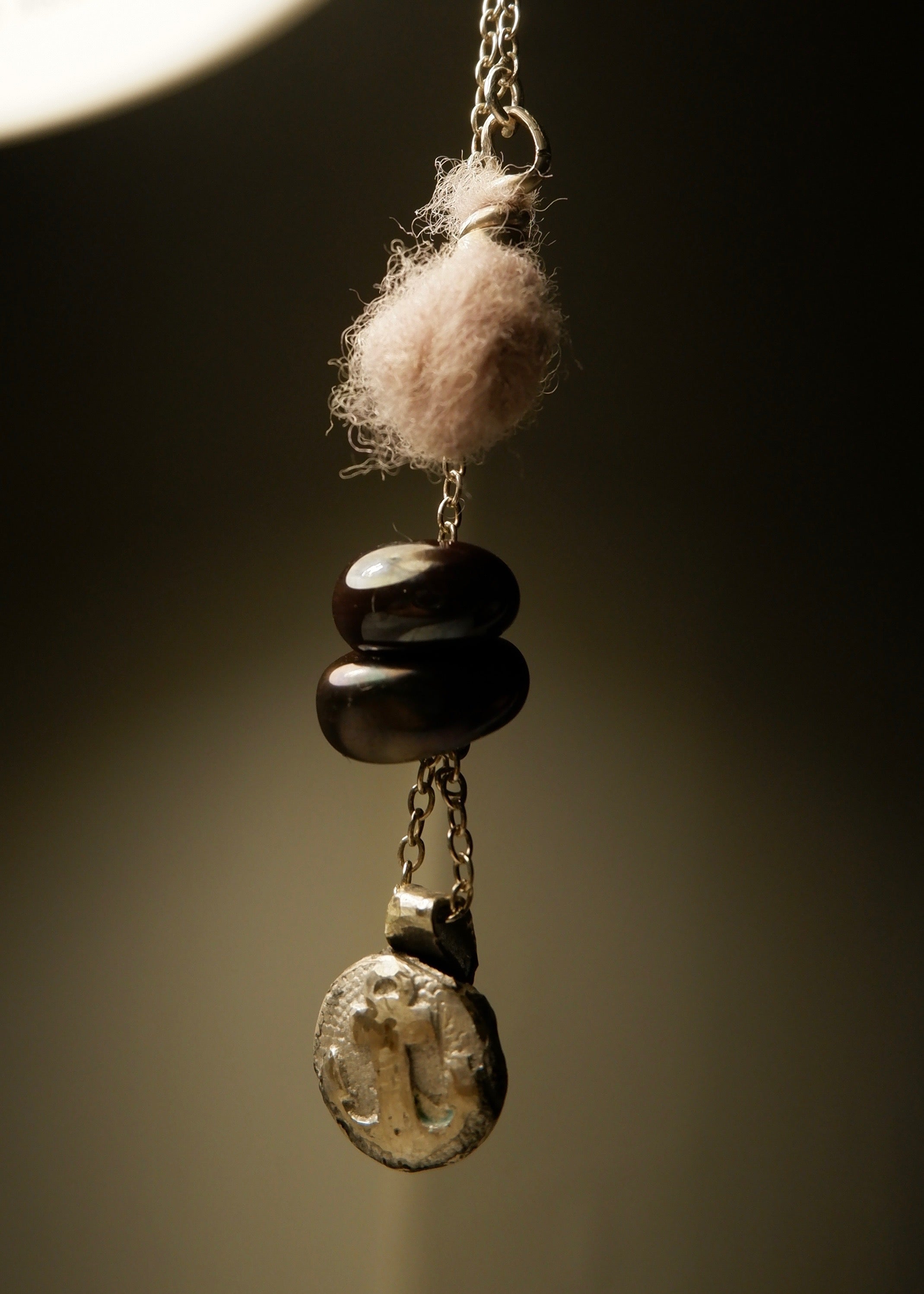A close-up of the Sunk • Ash Espinoza x Thyne Studio necklace by Thyne Goods, featuring a round textured charm, stacked dark beads, and a small wool ball on a recycled sterling silver chain against a neutral background.