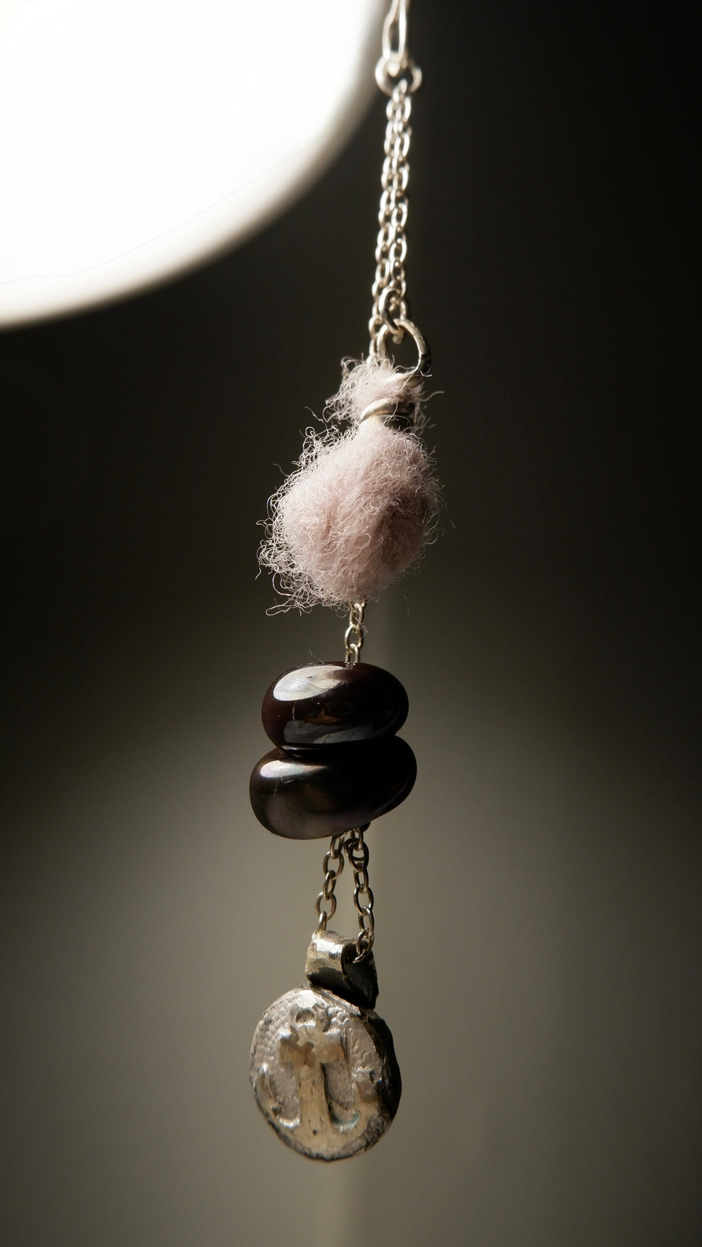 A close-up of the Sunk pendant by Ash Espinoza x Thyne Studio for Thyne Goods, featuring a pink wool ball, three dark oval beads, and a recycled sterling silver charm on a silver chain, crafted in Hyde Park, NY.