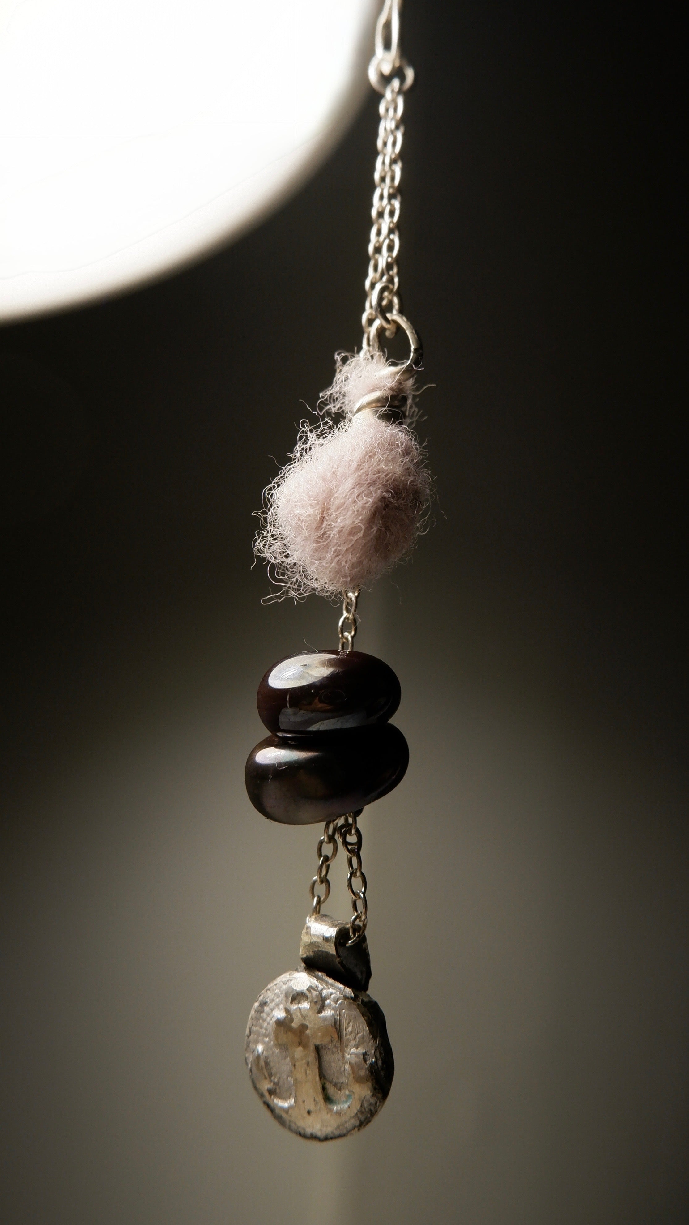A close-up of the Sunk pendant by Ash Espinoza x Thyne Studio for Thyne Goods, featuring a pink wool ball, three dark oval beads, and a recycled sterling silver charm on a silver chain, crafted in Hyde Park, NY.