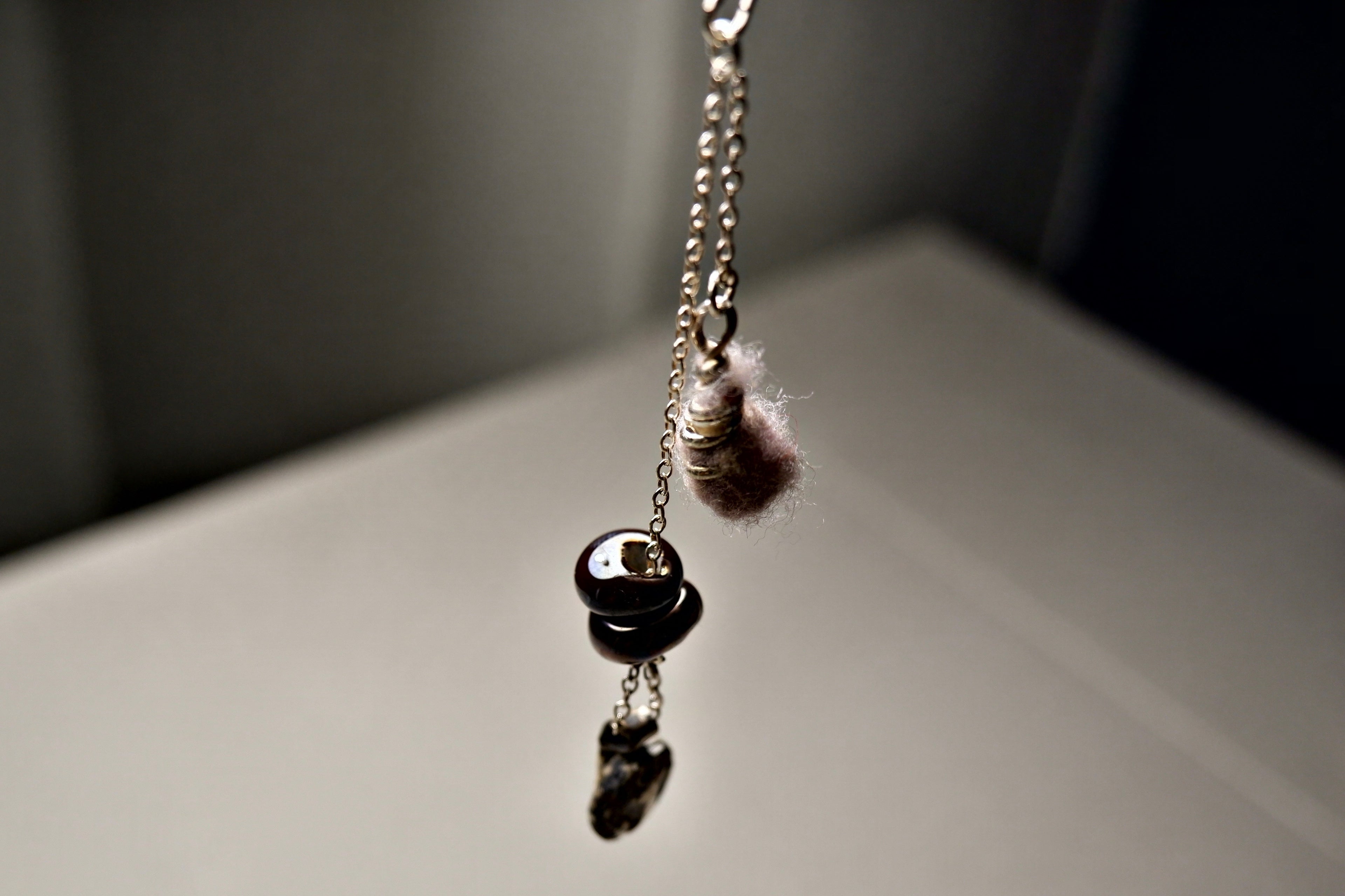 A close-up of the Sunk pendant necklace by Thyne Goods, designed with recycled sterling silver, a metallic bead, and a fuzzy pink charm on a delicate chain, all reflecting on a smooth surface.