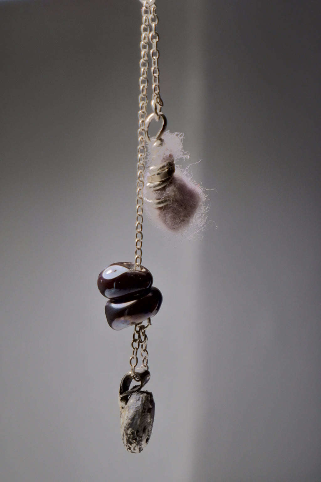 The Sunk necklace by Thyne Goods (Ash Espinoza x Thyne Studio) features a gold chain with unique pendants: a pink fuzzy piece, glass art beads, and a grey stone, each hanging separately on a plain grey background. Inspired in Hyde Park, NY.