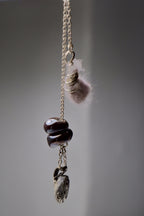 The Sunk necklace by Thyne Goods (Ash Espinoza x Thyne Studio) features a gold chain with unique pendants: a pink fuzzy piece, glass art beads, and a grey stone, each hanging separately on a plain grey background. Inspired in Hyde Park, NY.