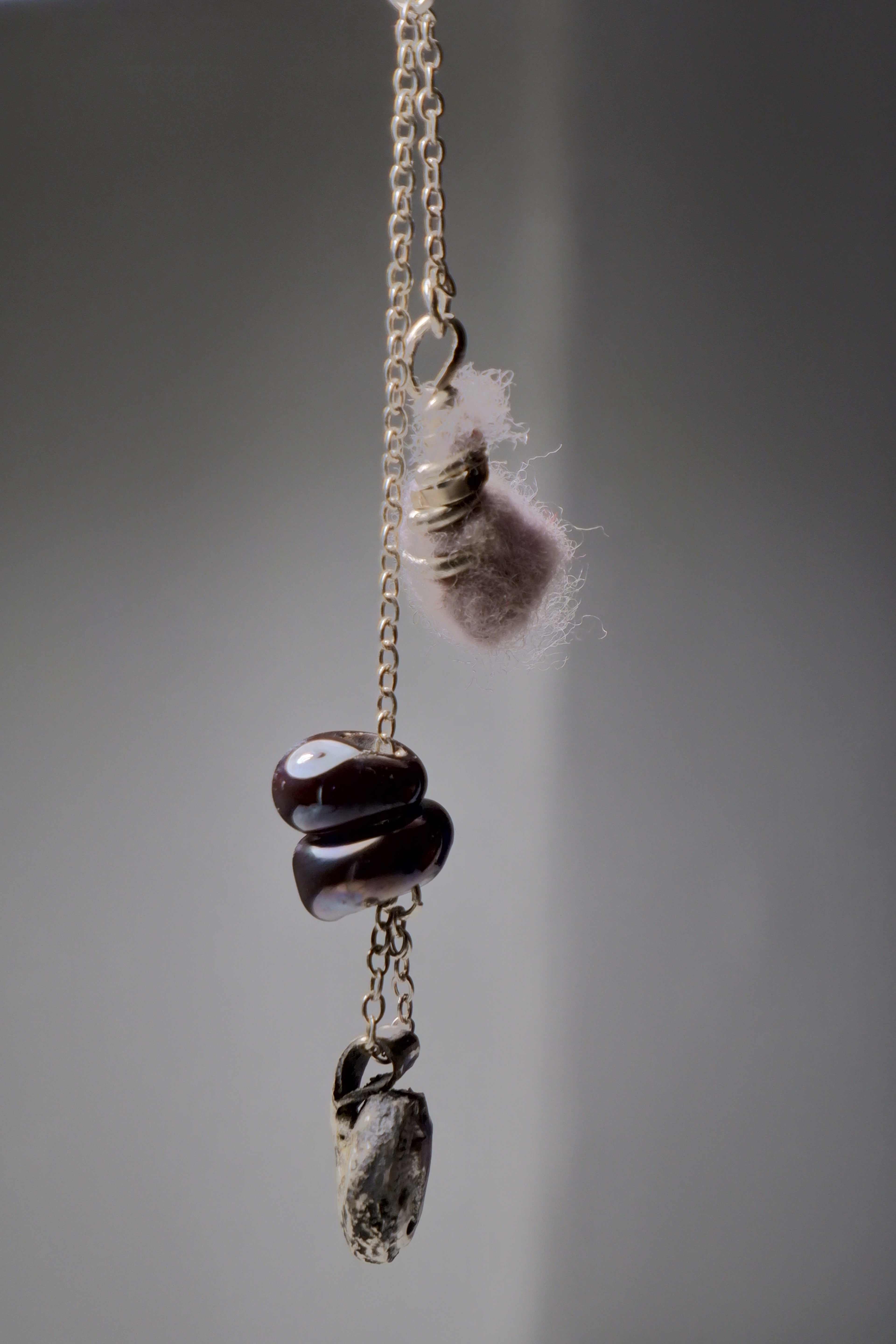 The Sunk necklace by Thyne Goods (Ash Espinoza x Thyne Studio) features a gold chain with unique pendants: a pink fuzzy piece, glass art beads, and a grey stone, each hanging separately on a plain grey background. Inspired in Hyde Park, NY.