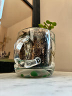 A small plant grows in a Thyne Goods Sam Lien • Handblown Glass Mug 1, filled with soil and water, on a marble surface. The clear handle and green dots add charm, with a blurred window and mirror in the background.