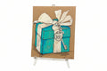 A painting titled "Rachel Nelson • Tiffany" by Thyne Goods features a blue gift box with a white bow and a Tiffany & Co. New York tag, displayed on a clear easel—a great pick for fans of Rachel Nelson art and neon flower paintings.