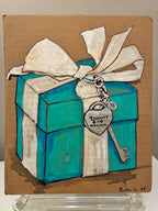 Rachel Nelson • Tiffany by Thyne Goods features a blue gift box with a white ribbon and silver key tag reading "Tiffany & Co New York," painted on brown paper and displayed on a clear stand.