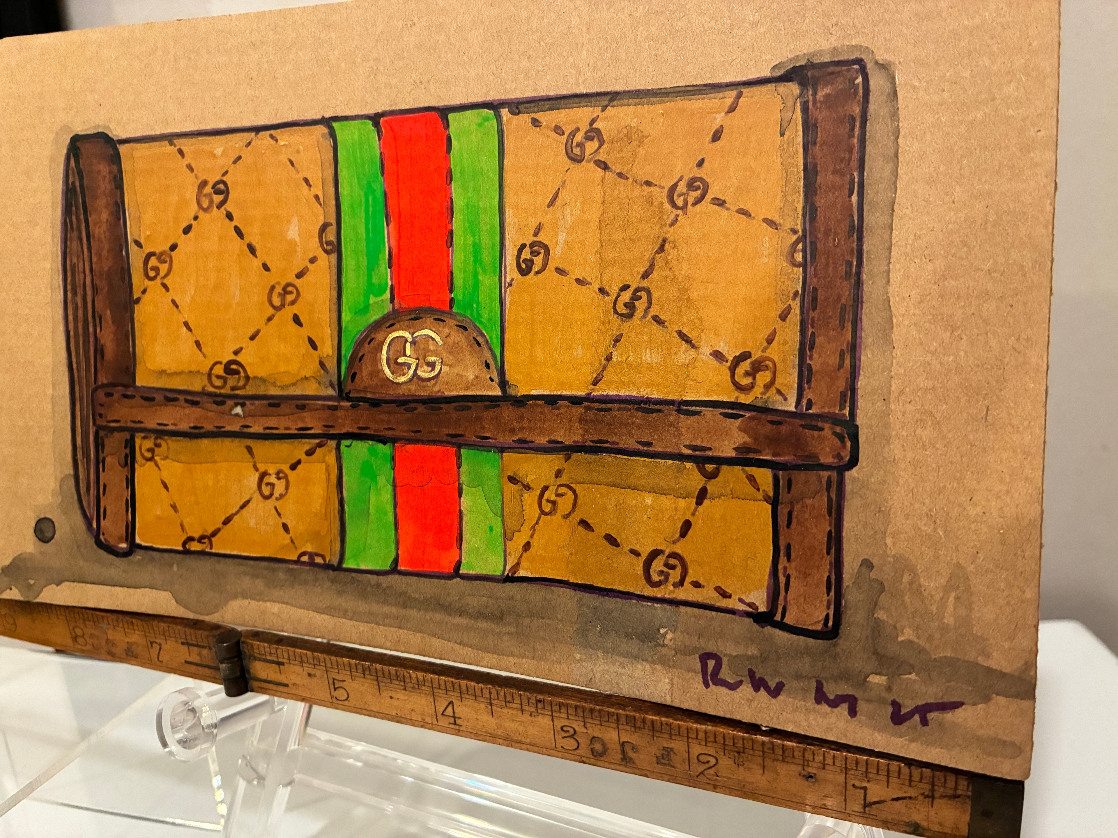 Rachel Nelson • Guch by Thyne Goods is a hand-drawn illustration of a designer-style clutch with a brown body, interlocking G patterns, green and red stripe, and brown accents, displayed on a wooden ruler stand.